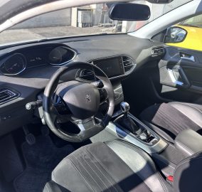 Interior view from the driver’s seat showing the steering wheel, dashboard gauges, and center touchscreen infotainment, with a gear lever in the foreground.