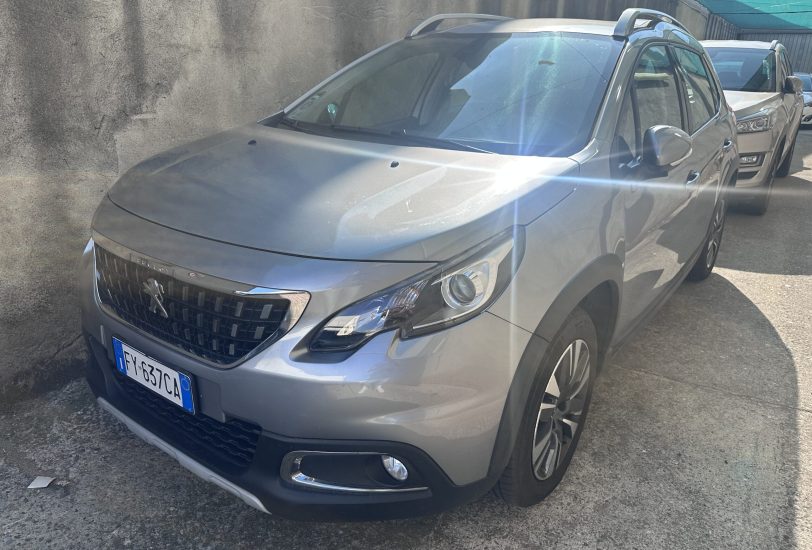 Front-right angle of a silver SUV parked in a narrow lot beside a concrete wall, with sun glare across the windshield and hood.