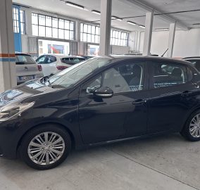 Indoor car showroom with multiple parked cars and large industrial windows in the background, white walls, and blue panels on the left side.