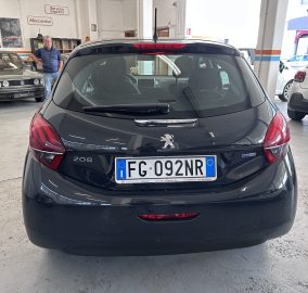 Rear view of a dark Peugeot 208 in a car workshop, with a man in a blue vest standing near an older car on the left (Italy plates).