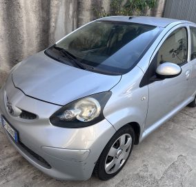 Front-left view of a silver compact car parked along a textured concrete wall. Shows the windshield, wipers, driver’s door, and side mirror.