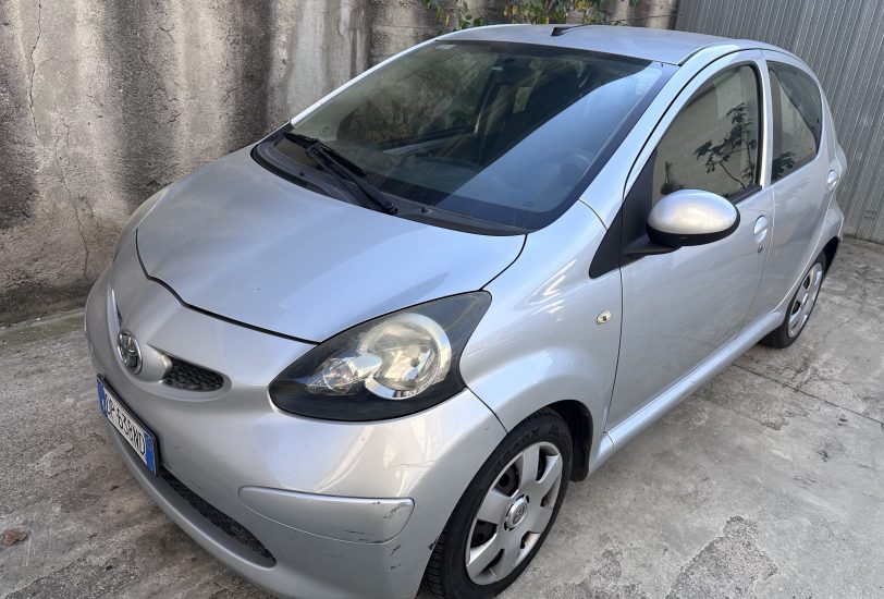 Front-left view of a silver compact car parked along a textured concrete wall. Shows the windshield, wipers, driver’s door, and side mirror.