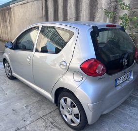 Rear-right side view of a silver Toyota hatchback parked on a concrete driveway beside a tall, weathered wall with vines growing along it.