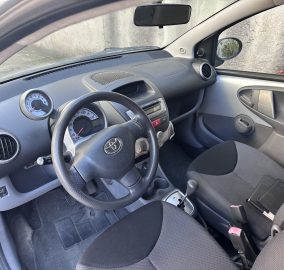 Front-of-car interior: steering wheel, dashboard, and driver’s seat with a side mirror visible through the window.