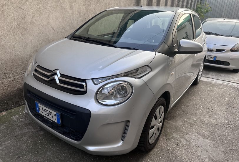 Front left of a silver Citroën car parked beside a beige wall, with another white car in the background.