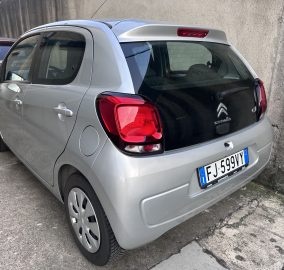 Rear view of a silver Citroën hatchback parked beside a concrete wall, with another car visible to the left.