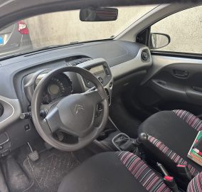 Driver’s seat view inside a compact car showing the steering wheel, dashboard, gear lever, and striped cloth seats with a nearby cup holder.