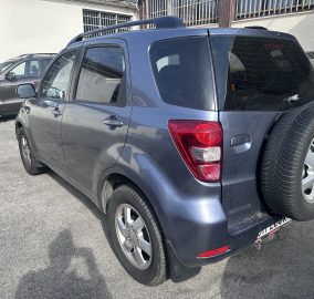 Rear-quarter view of a blue SUV in a parking lot, with a spare tire mounted on its rear door and another car beside it under a building with barred windows; daytime.