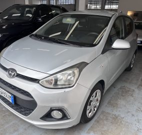 Front three-quarter view of a silver Hyundai in an indoor parking area with other cars nearby and polished concrete floor. Front bumper and grille visible, license plate partially shown.