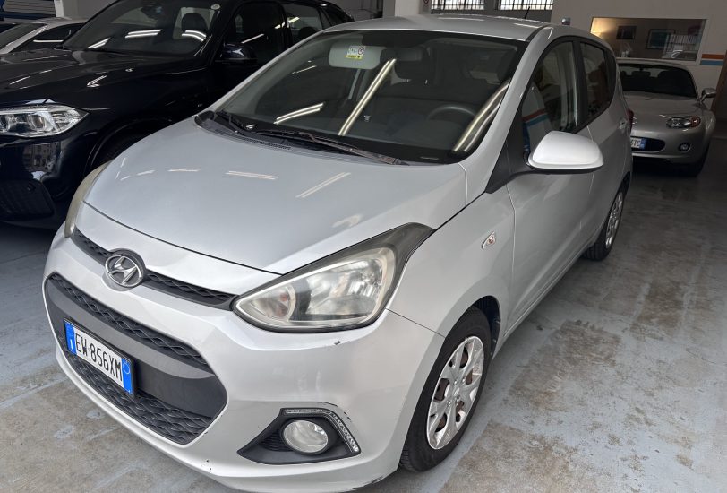 Front three-quarter view of a silver Hyundai in an indoor parking area with other cars nearby and polished concrete floor. Front bumper and grille visible, license plate partially shown.