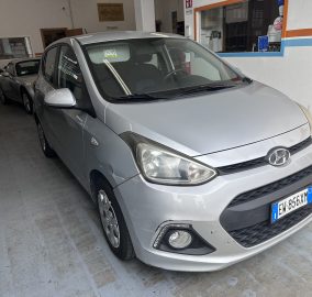 Three-quarter front view of a silver Hyundai minivan parked inside a workshop/garage with other cars and workshop equipment visible in the background