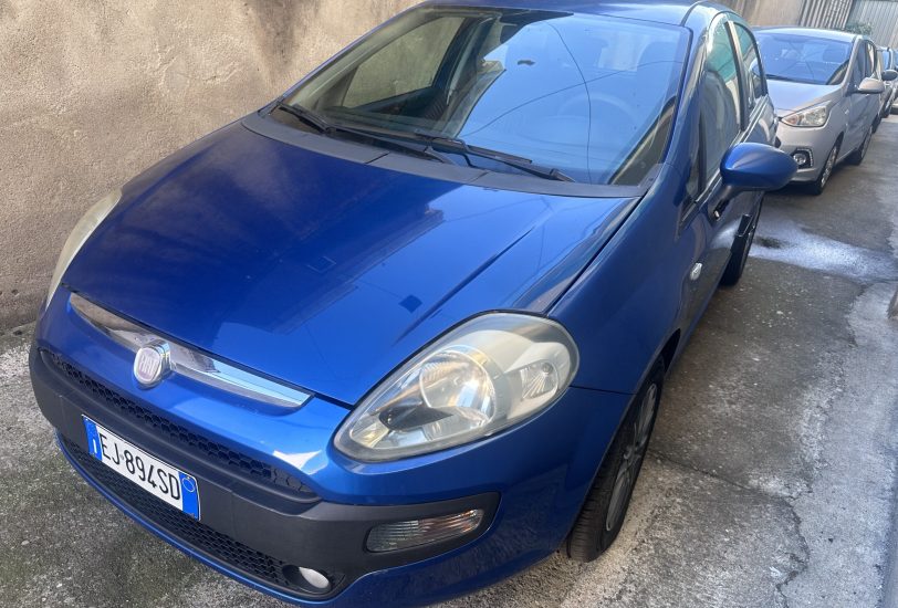 Blue Fiat car parked on a street with a tan wall in the background, other cars behind it partially visible.