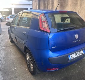 Blue Fiat Punto hatchback parked along a narrow alley, showing the rear and passenger side against a concrete wall behind it.