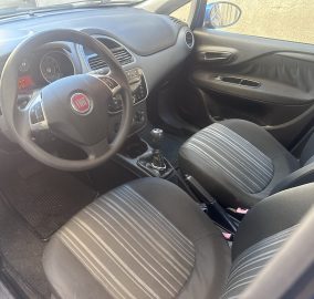 Front interior of a Fiat car showing steering wheel with logo, dashboard, center console, manual gear shifter, and striped seats context.
