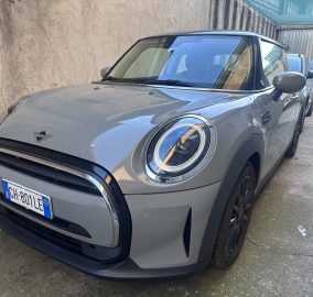 Front-left view of a gray Mini Cooper parked in a narrow lot, with a beige wall on the left and other cars in line behind it.