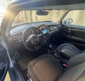 Interior of a MINI: driver and front passenger seats with a circular center display and steering wheel, sunlit dashboard.