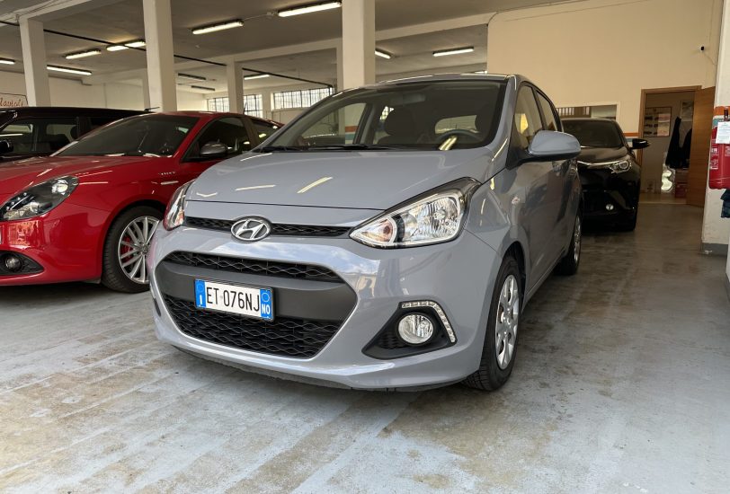 Grey Hyundai hatch in a bright indoor showroom or garage, with a red car parked to its left and other vehicles in background.