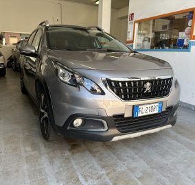 Gray Peugeot SUV parked indoors in a garage, with a white car to the left and a service counter window on the wall behind.