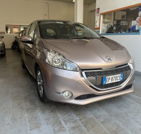 Front three-quarter view of a beige Peugeot car parked in an indoor garage beside a service counter with blue/orange wall stripes.