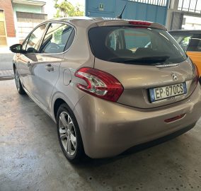 Beige Peugeot hatchback parked in a garage, shown from the rear three-quarter view with an orange car beside it.