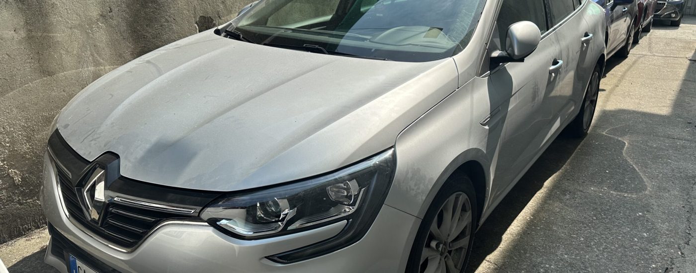 Front-left view of a silver SUV parked along a concrete wall with several cars lined up behind it under a green canopy.