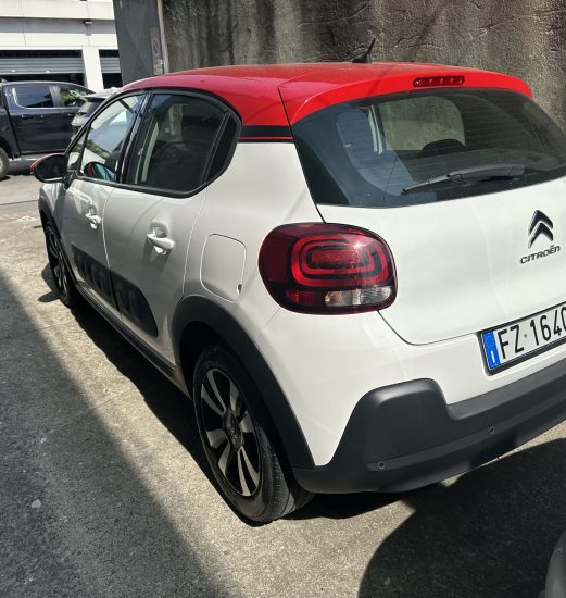 Rear three-quarter view of a white Citroën SUV with a red roof, parked in a lot near a textured wall. The rear lights and windshield wiper are visible, along with a blue European license plate.