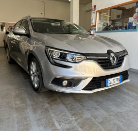Silver Renault SUV parked inside a garage showroom, front-right view with another car in the background on the left side, and a service window on the right wall.