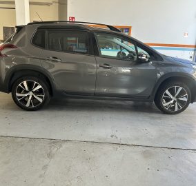 Gray SUV inside a garage, showing alloy wheels and roof rails, with another car in the background and a fire extinguisher on the wall behind.