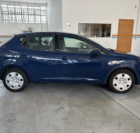 Blue hatchback parked indoors in a garage showroom with another car visible on the left and orange/blue wall stripes behind it