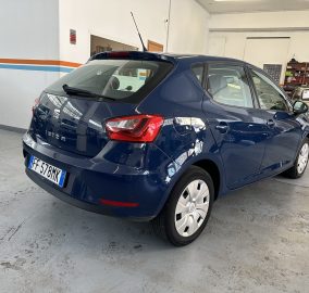 Rear three-quarter view of a blue SEAT Ibiza parked in a garage, with a beige vintage car and shelves visible in the background.