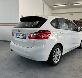 White BMW 218i SUV parked in an indoor garage, seen from the rear with a vintage car in the background on the right side.