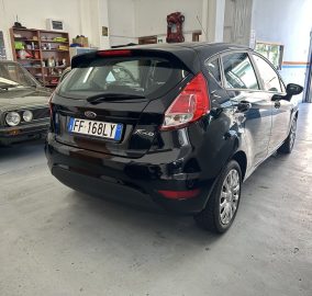 Black Ford Fiesta hatchback parked in a service workshop, with a black SUV nearby and toolshelves in the background.”Note: include correct punctuation only where needed.