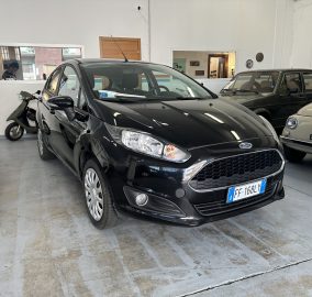 Indoor car showroom with a black Ford in the foreground and other vehicles visible along the sides, including a beige Fiat on the right and a scooter on the left window area.