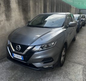 Line of parked cars along a narrow alley with a tall concrete wall on the left, gray SUV in foreground and more cars behind, green netting overhead.