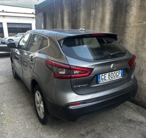 Gray Nissan Qashqai parked in a narrow lot beside a tall concrete wall, rear view with visible Italian license plate GE 800GP