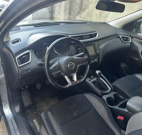 Driver's-side view of a Nissan car interior: steering wheel, dashboard, center touchscreen, gear stick, and black seats.