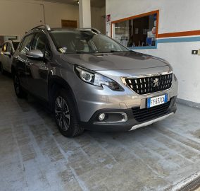 Silver Peugeot SUV parked inside a garage or workshop, with other vehicles visible in the background and orange-blue wall stripes nearby.