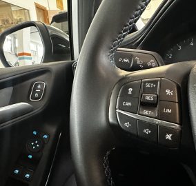 Close-up of a car steering wheel and control stalk with cruise-control buttons, plus window/mirror controls and dashboard visible in a garage interior.