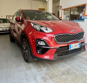 Red Kia SUV parked in a garage or showroom bay with a white wall and service window in the background, front grille visible.