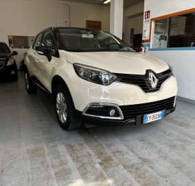 White Renault SUV parked indoors in a showroom, with a black car on the left and orange-blue wall trim in the background.