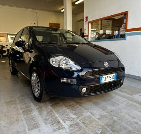 Black Fiat hatchback parked inside a garage service bay, with a blue car and a scooter visible in the background.