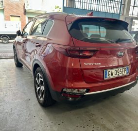 Red Kia Sportage SUV shown from the rear-side, parked in a garage or showroom with another car beside it and blue walls in the background