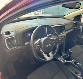 Inside car cabin showing steering wheel with Kia logo, dashboard, center console, and gear shifter; red exterior visible at left edge.