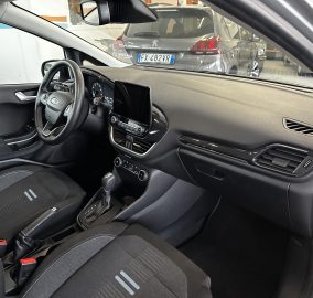Interior of a car cockpit seen from the passenger side: steering wheel, dashboard, center touchscreen, and gear shifter; dark seats with white stitching.