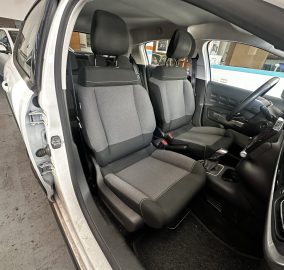 Open car door reveals the front interior: two black fabric seats, steering wheel and dashboard on the right, in a showroom with other vehicles outside the window.