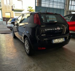 Rear three-quarter view of a black Fiat Punto parked in an indoor garage, with a red SUV to the right and a pale blue wall in the background.